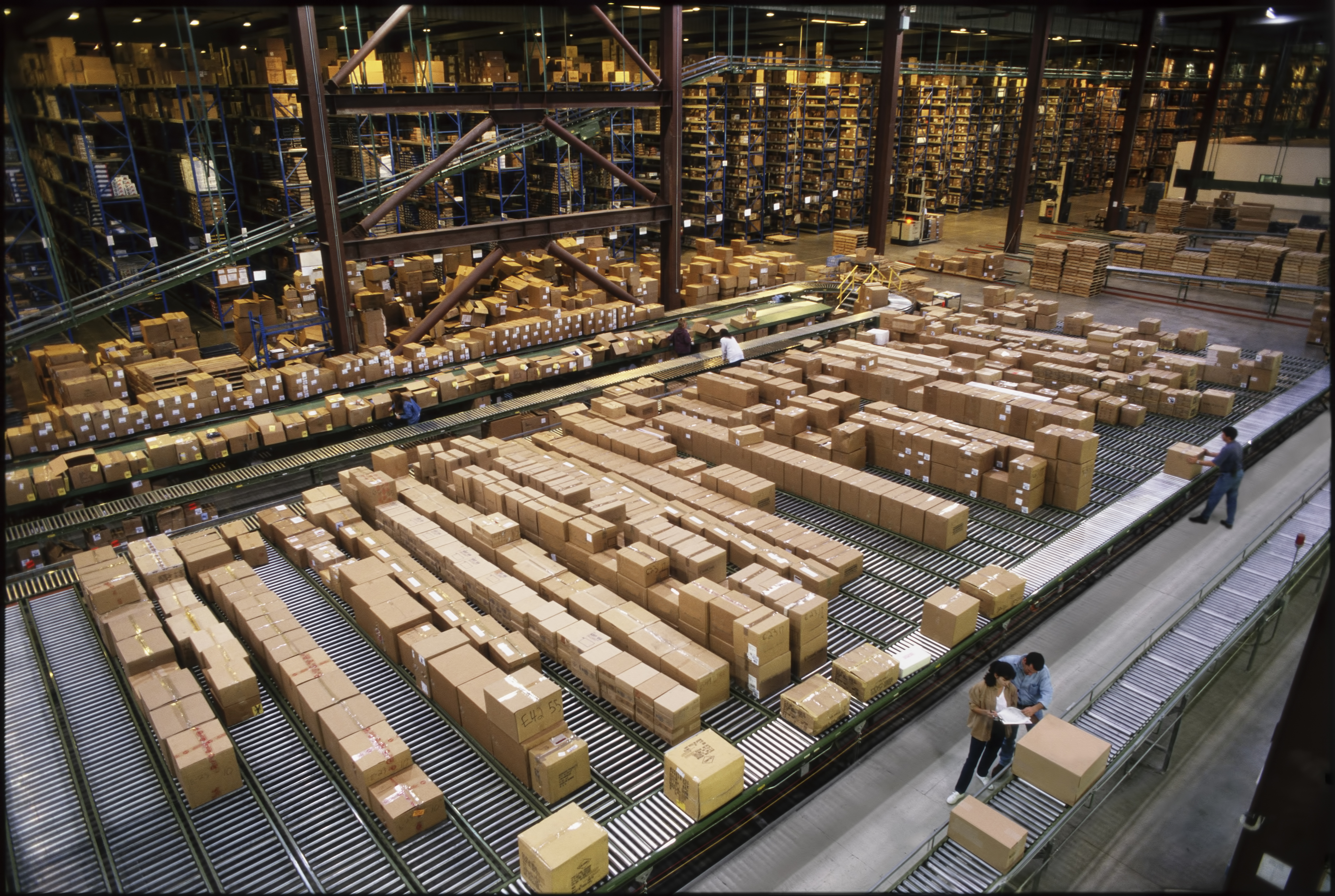 Vue d'ensemble d'un grand entrepôt de distribution industrielle stockant des produits dans des boîtes en carton sur des tapis roulants et des rayonnages.